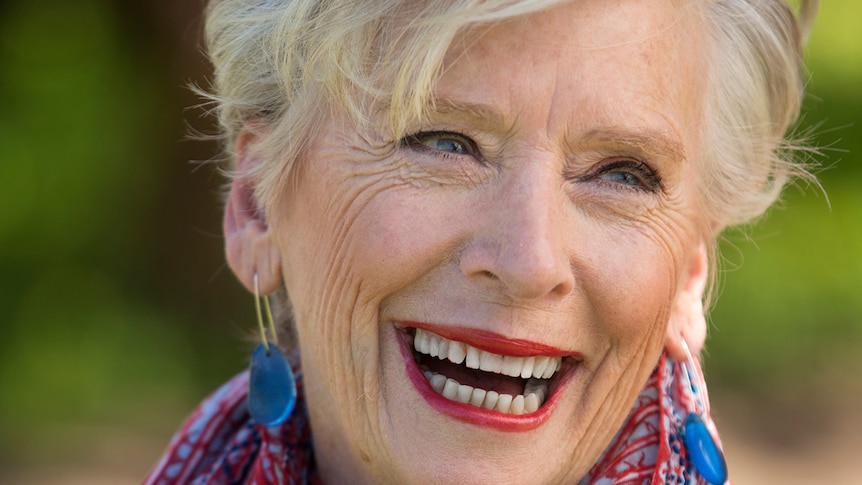 Maggie Beer has a big smile. She has grey hair, cut short, and is wearing bright red lipstick and a scarf to match.