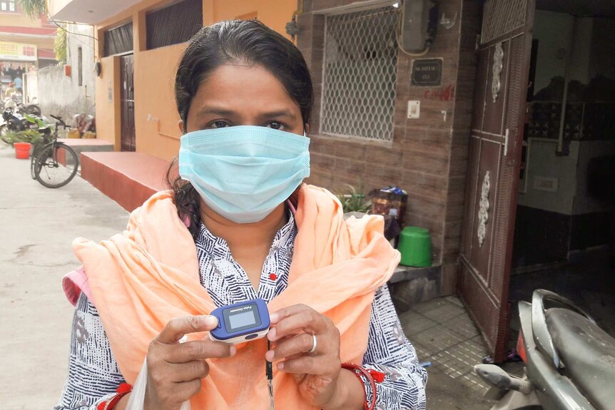 A woman with black hear wearing a mask and an orange scarf holds a device.