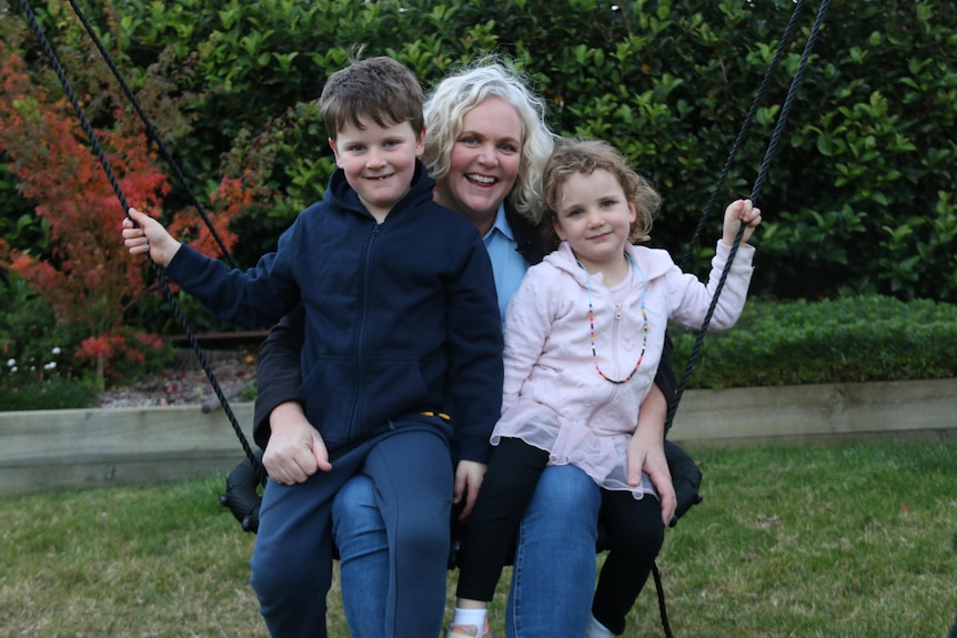 A woman sits on a swing in a suburban backyard, and her two children sit on her lap.