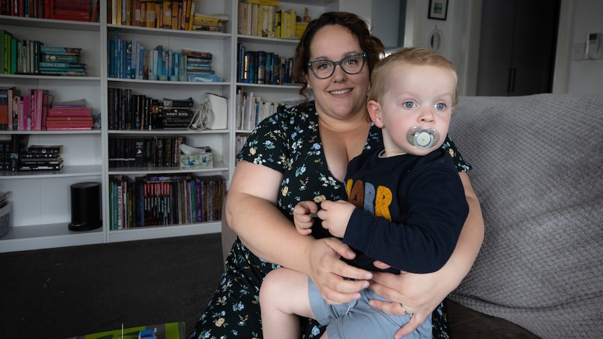 Emma Helks sits with two-year-old Theo on her lap, with a bookshelf in the background and toys on the floor