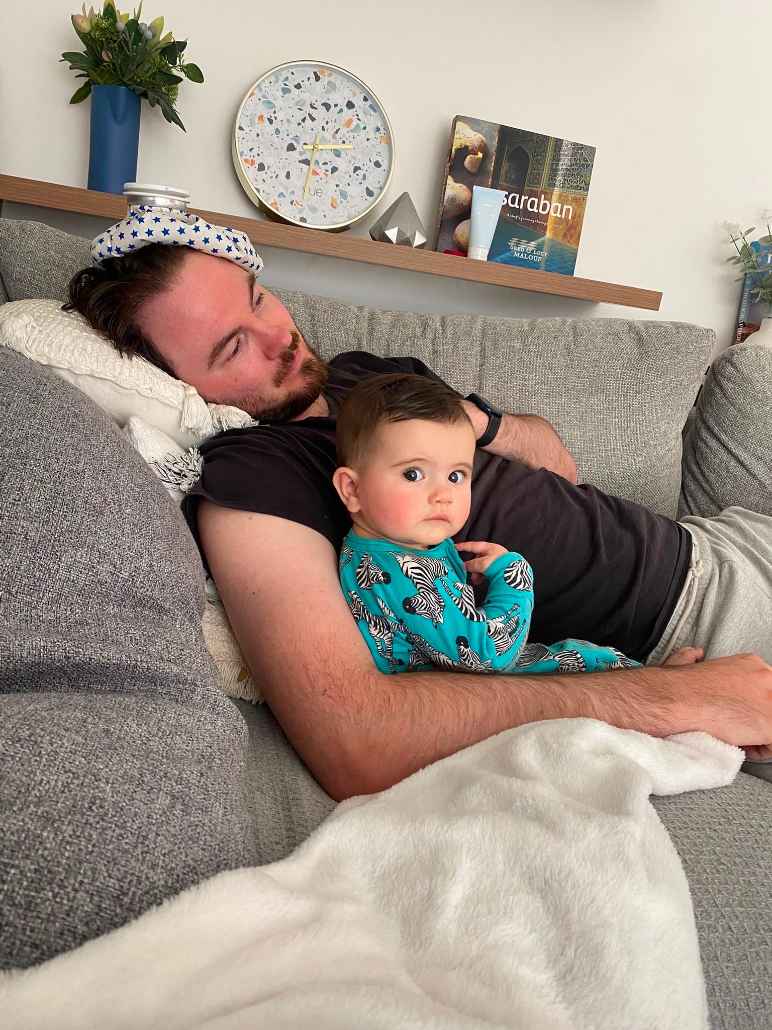 A man lying on a couch with an ice bag on his head and a baby next to him.