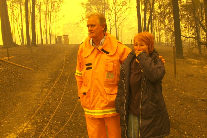Williams in yellow protective fire suit with arm around woman with hand over her mouth in shock, amid burnt bush and yellow sky.