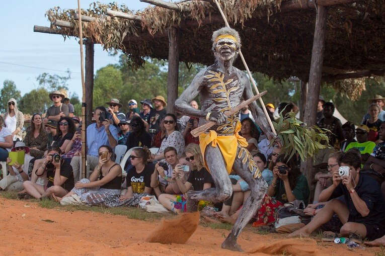 An Indigenous performer dances wearing white and yellow body paint as crowd looks on