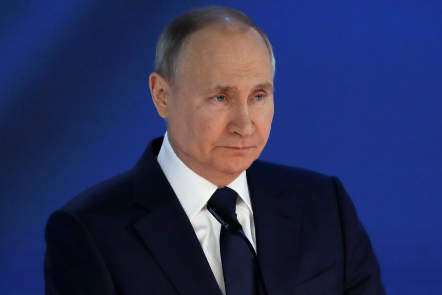 Vladimir Putin, standing in front of a blue background, looks stern.