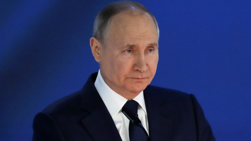 Vladimir Putin, standing in front of a blue background, looks stern.