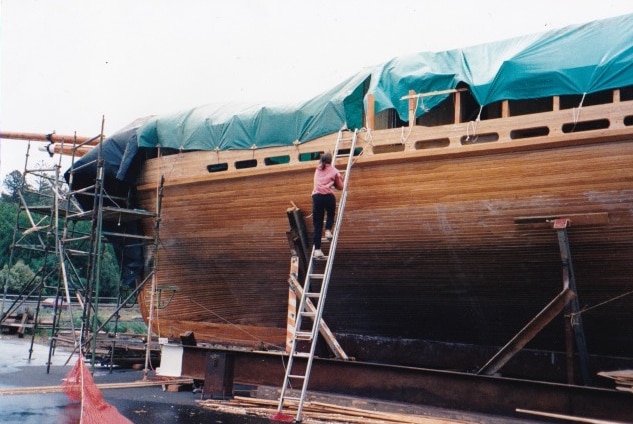 A large wooden boat being built with a woman on a ladder on the side of it