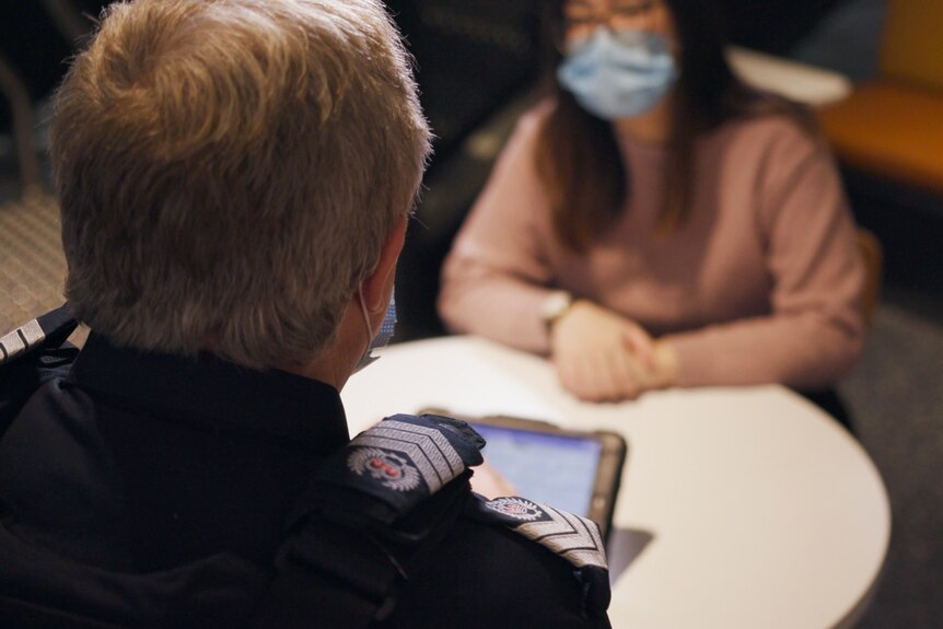 A male police officer holding an iPad talks to a woman sitting across a table