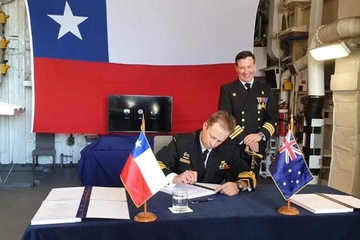 Two men stand behind a desk in naval gear. A Chilean flag is hanging behind them. One of the men smiles at the other.
