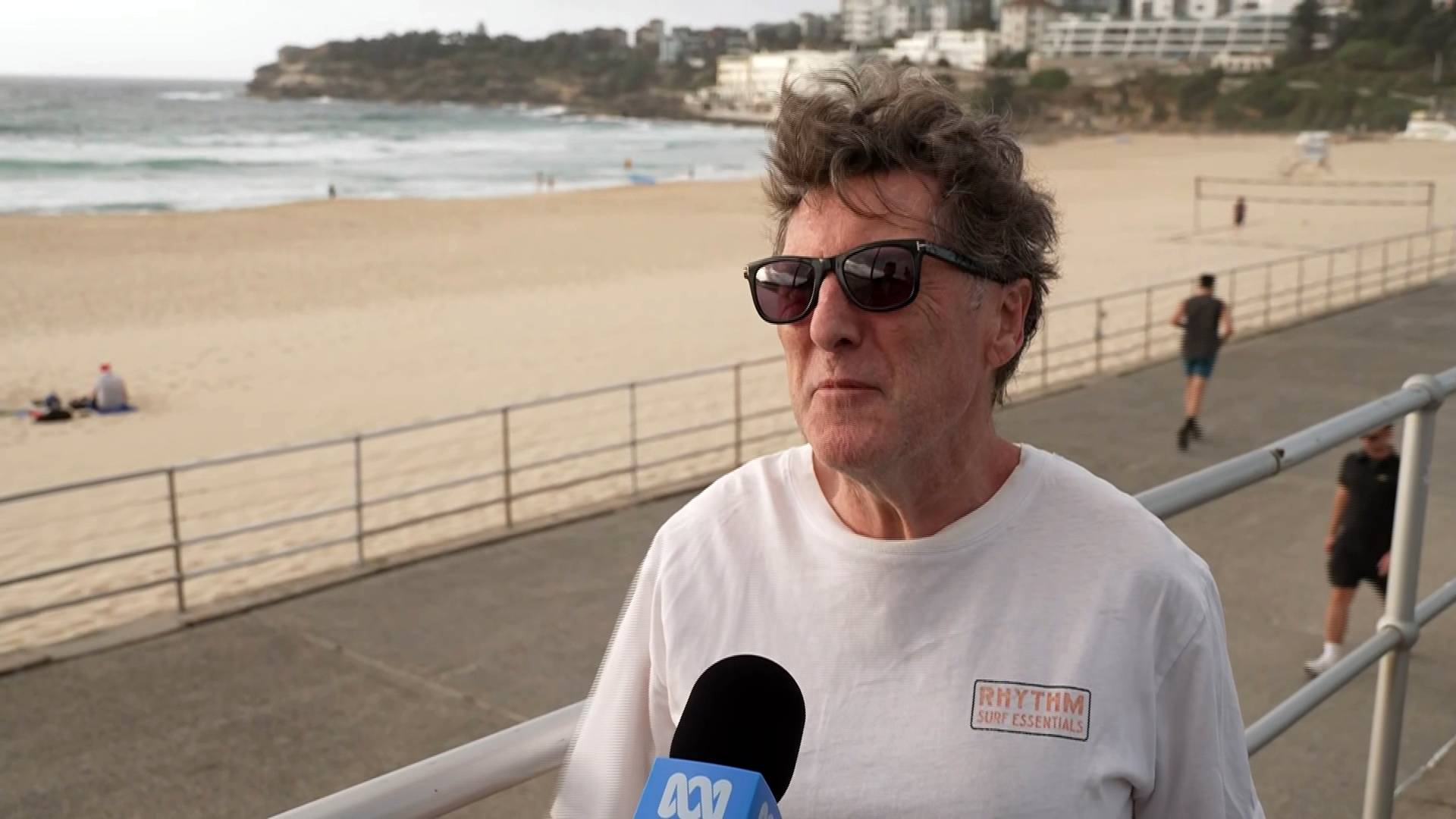 Um homem de óculos escuros conversando com um jornalista no calçadão de Bondi Beach.