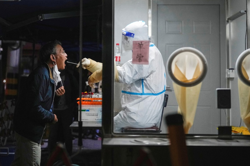 A man opens his mouth for a worker in a protective suit to swab it through a glass wall. 