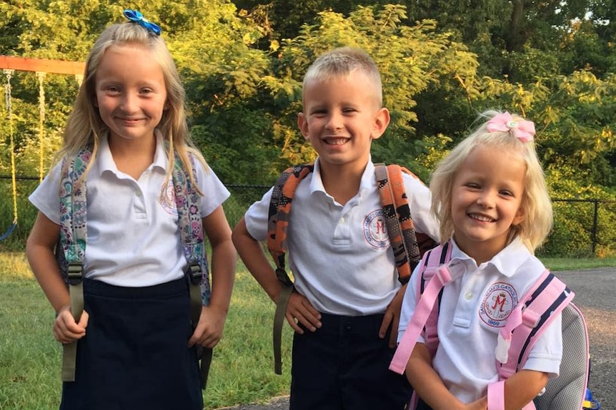 Three blonde children in school uniforms with backpacks on their backs