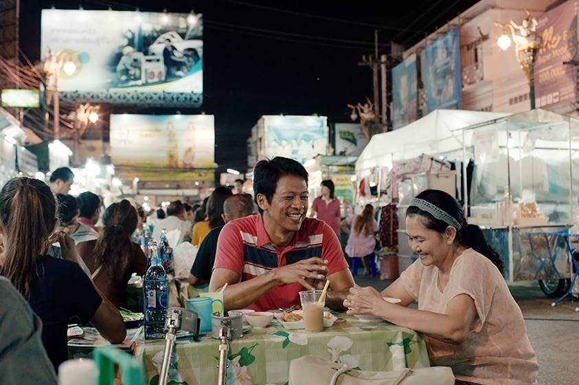 A middle aged man and woman sit at outdoor dining table in busy street at Thai night market while they eat and laugh.