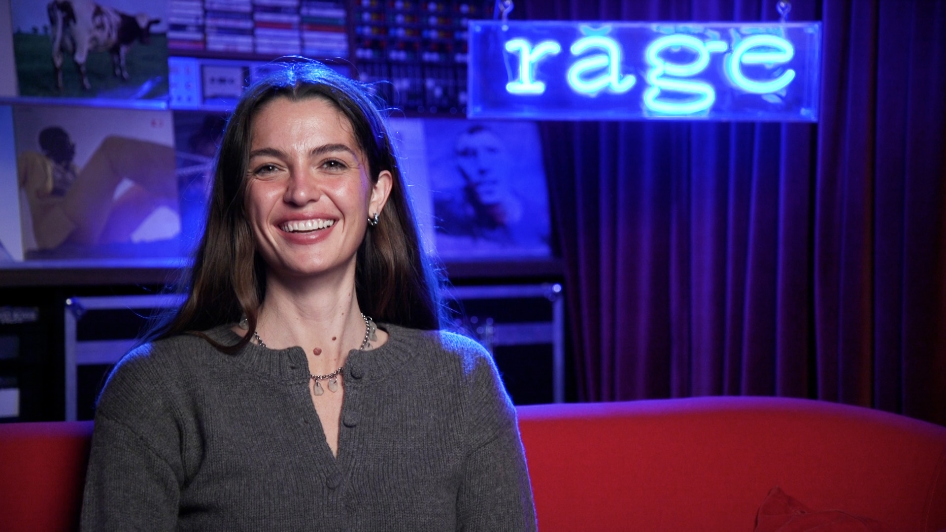 Kita Alexander sits smiling on a red couch in front of a neon blue sign that reads rage