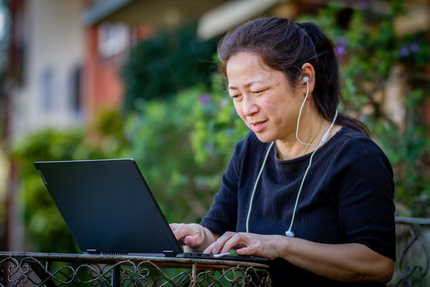 Lee Chong typing on a laptop on a small table near a garden outdoors, with a slightly serious expression on her face.