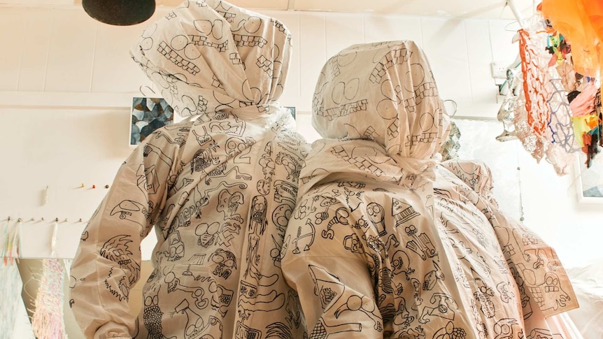 Two figures are clothes in full-body, hooded beige suits with black drawings all over them. You cannot see their faces.