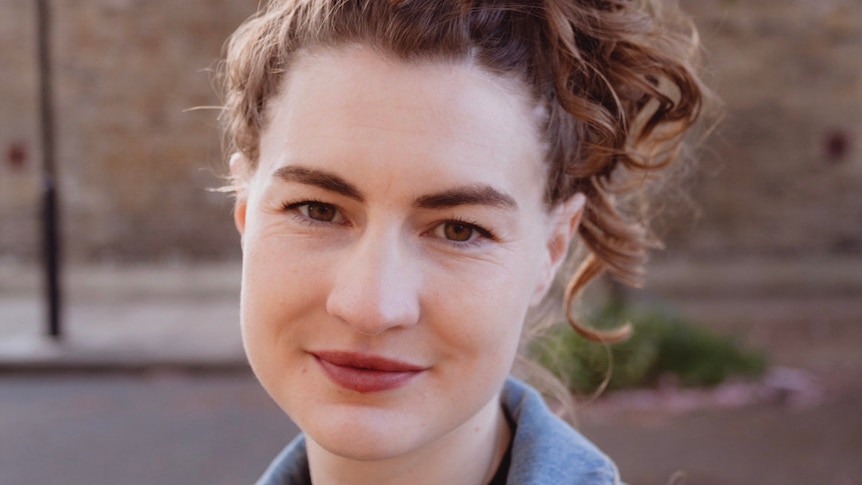 A close up photo of Lucia Osborne-Crowley, a woman with brown curly hair wearing brown lipstick and a blue shirt