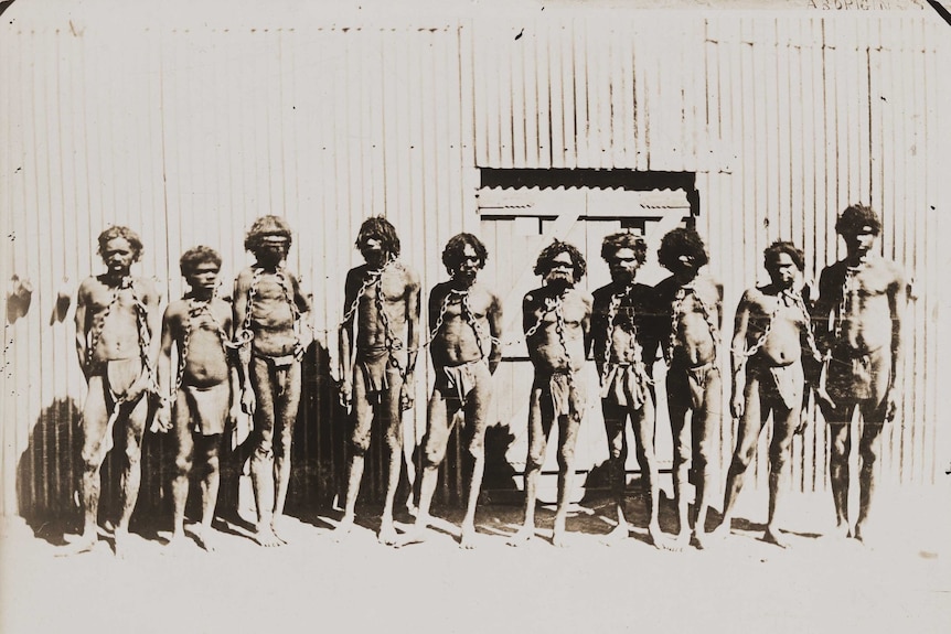Historical photo of four traditional Aboriginal prisoners, wearing only loin clothes, chained by the neck.