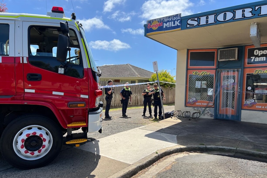 Police stand between a milk bar and parked fire truck putting up police tape.