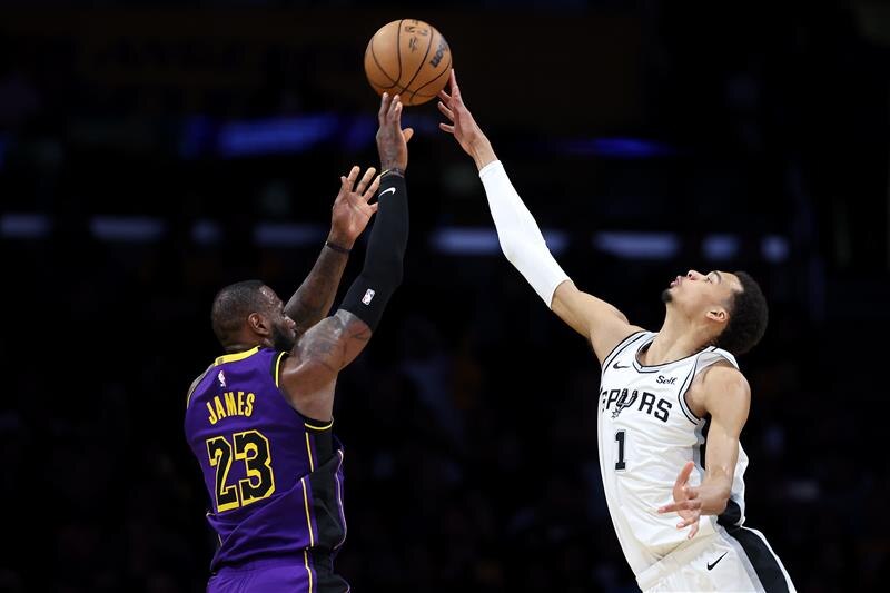 A tall player in white blocks a shot by a shorter player in purple during an NBA match.
