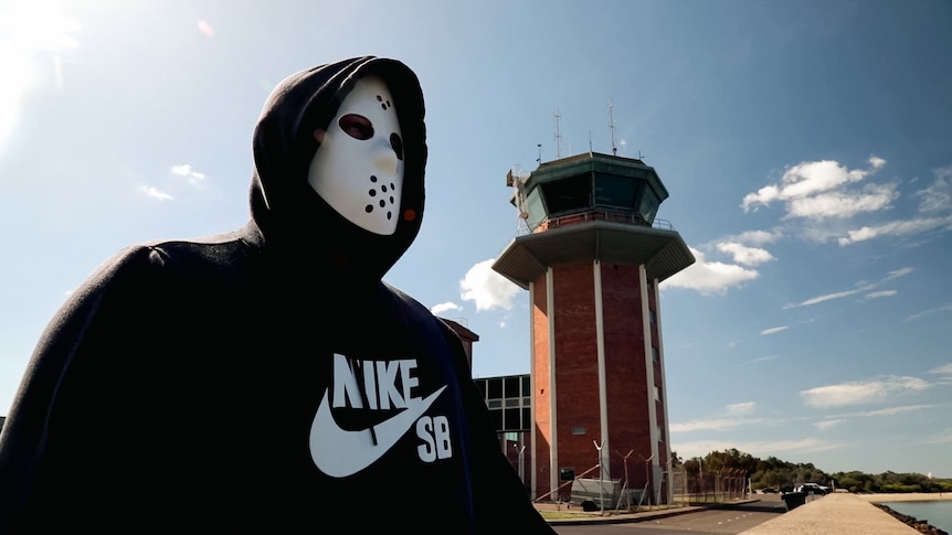 A man wearing a hockey mask and hoodie stands outside in front of an old airport control tower.