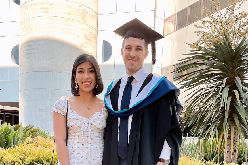 A man in a graduating gown and hat stands next to a woman in a dress, smiling.