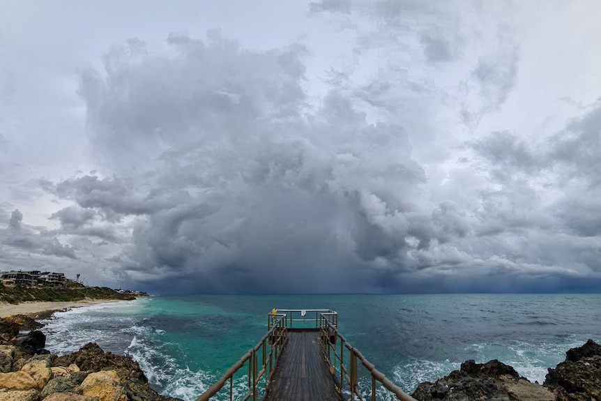 Dark storm clouds over the Perth coastline with a small jetty in the foreground and houses along the water on the left.