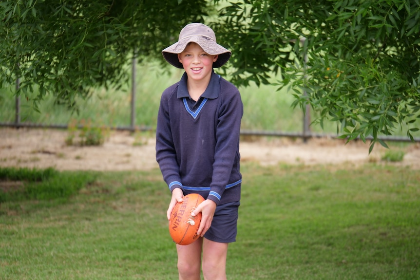 A boy in school uniform wears a hat, holds a football and smiles at the camera.