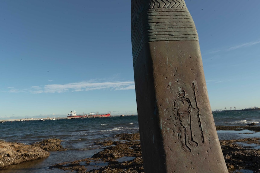 The figure of a man holding a spear carved into part of a bronze sculpture by the sea. In the background ships can be seen.