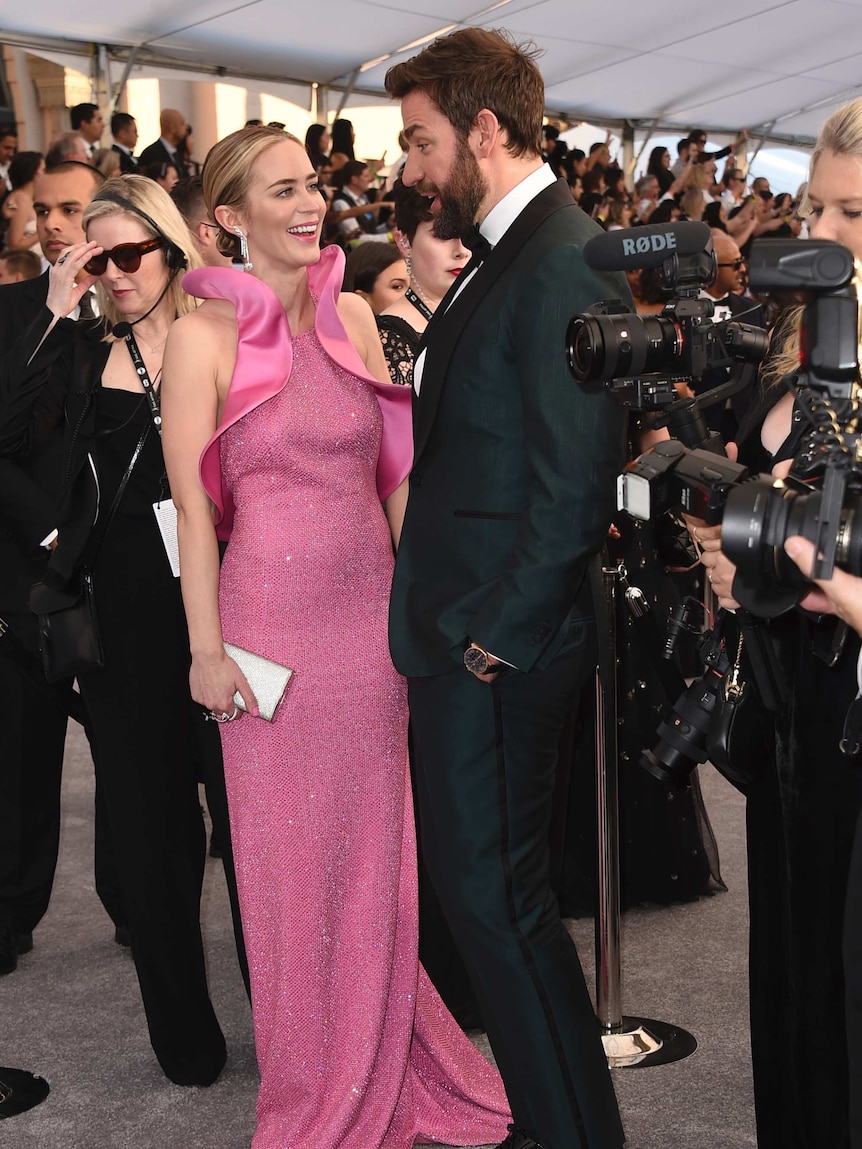 Emily Blunt wears pink gown as she smiles up at husband John Krasinski wearing emerald tuxedo while surrounded by photographers.