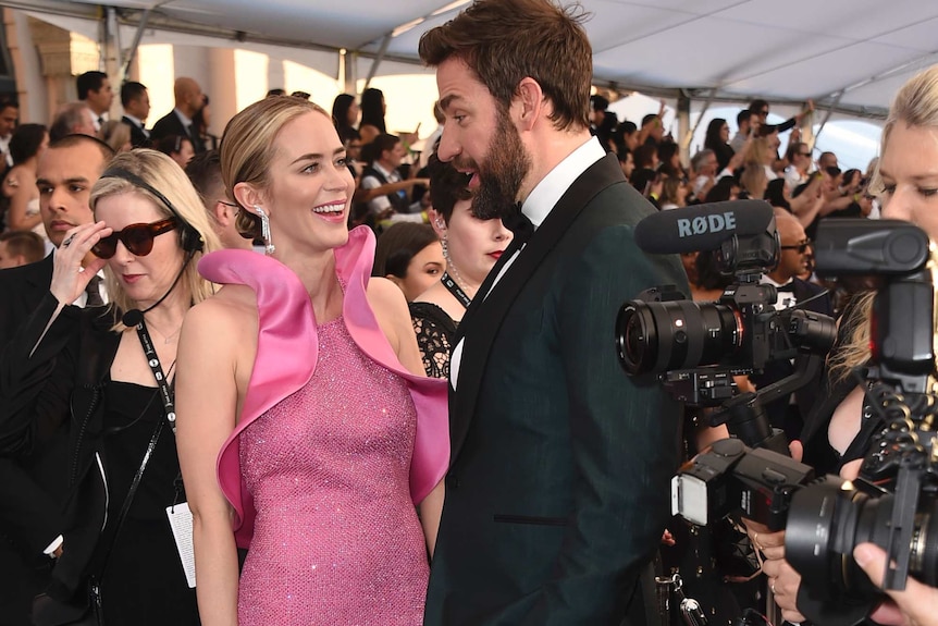 Emily Blunt wears pink gown as she smiles up at husband John Krasinski wearing emerald tuxedo while surrounded by photographers.