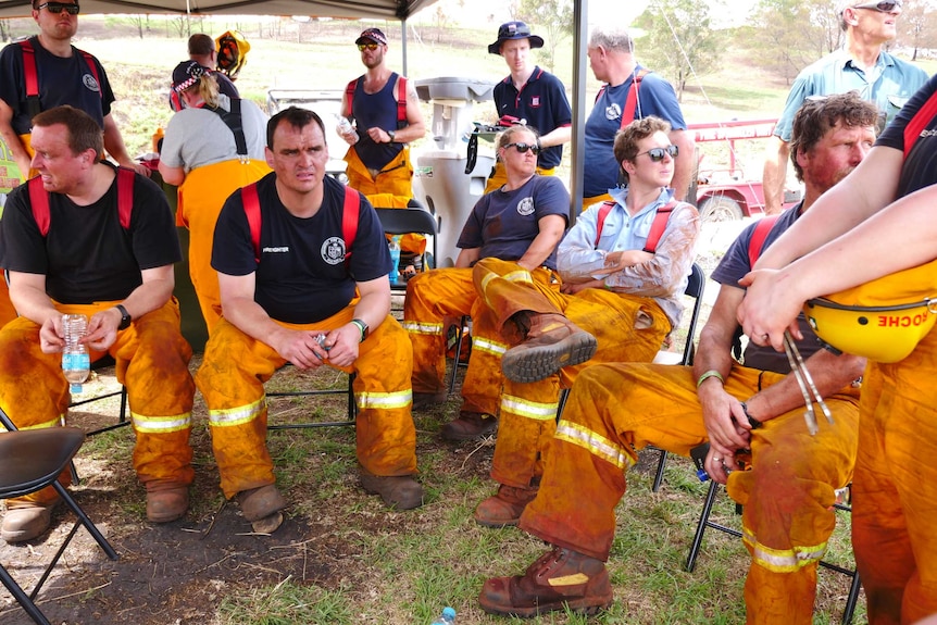 A group of men in high-vis firefighting uniforms take a break sitting under an outdoor marquee shade.