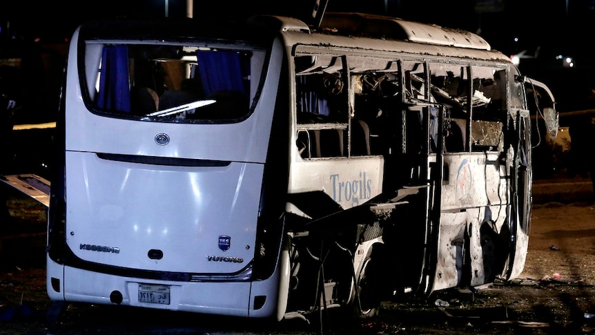 A destroyed tourist bus sits on a the side of a road with its side windows and doors blown off by a bomb.