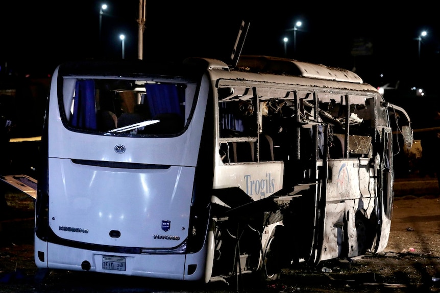A destroyed tourist bus sits on a the side of a road with its side windows and doors blown off by a bomb.