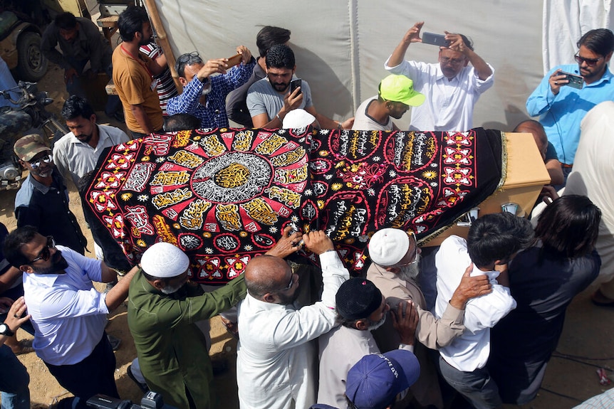 A group of men carrying a casket draped with a colourful red, yellow and black cloth, while others record the event with phones