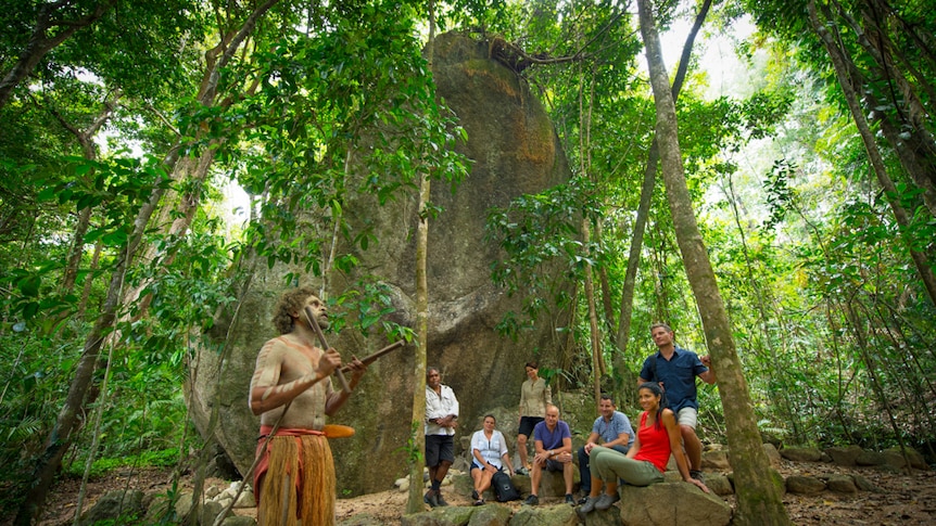 An Indigenous man beating clap sticks, with a group of tourists watching on in the rainforest.