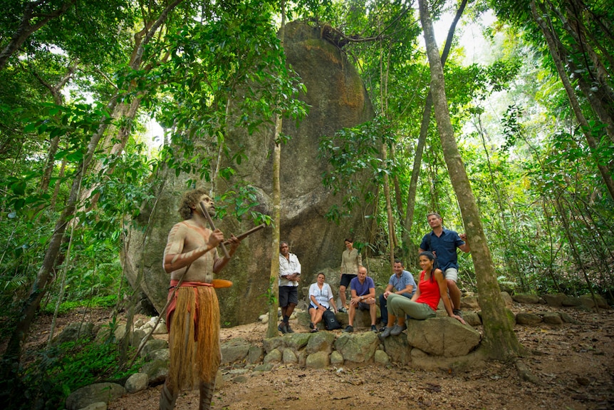 An Indigenous man beating clap sticks, with a group of tourists watching on in the rainforest.