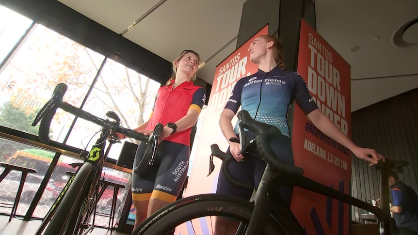 Two women in cycling outfit holding on to their bikes.