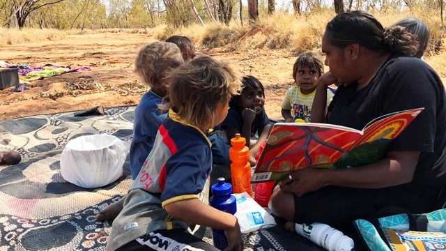A group of Aboriginal children are read to by an Aboriginal woman in the bush.