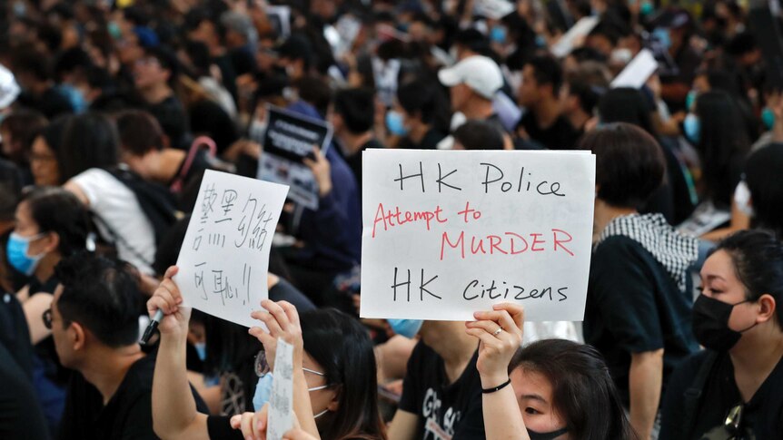 A close up of two women in large crowd holding up sings, one says "HK police attempt to murder HK citizens"