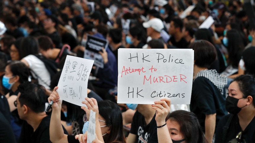 A close up of two women in large crowd holding up sings, one says "HK police attempt to murder HK citizens"