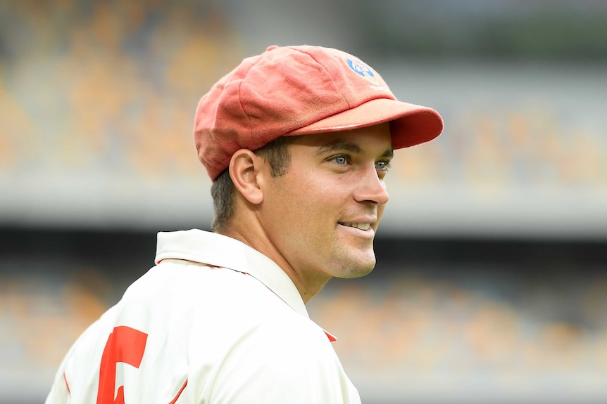 A male cricket player wearing a red cap smiles as he looks to his right.