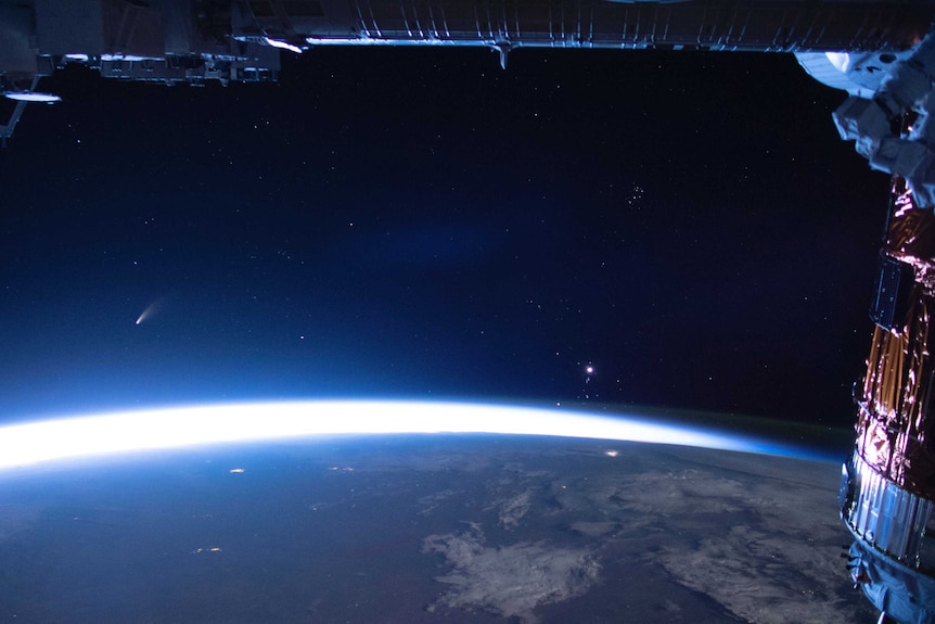 A portion of the International Space Station is seen above Earth with Comet Neowise trailing above the atmosphere