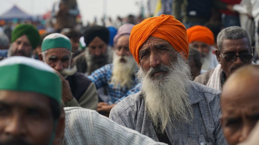 a man wearing orange headwear sits among a crowd of seated protesting farmers