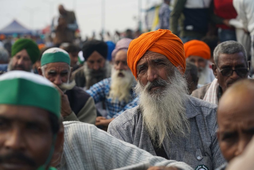 a man wearing orange headwear sits among a crowd of seated protesting farmers