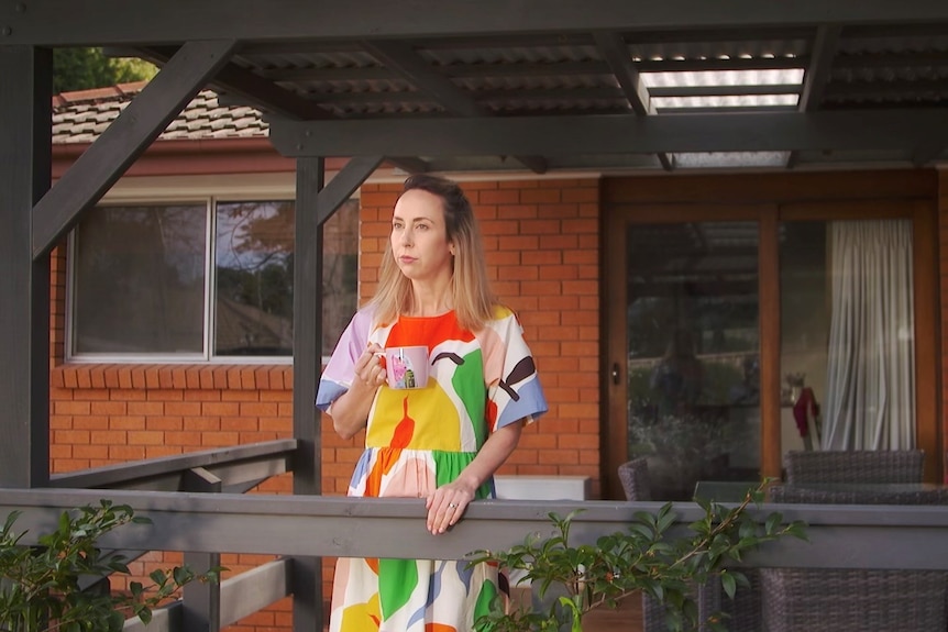 A lady in a colourful dress stands on her porch with a coffee mug staring into the middle distance 