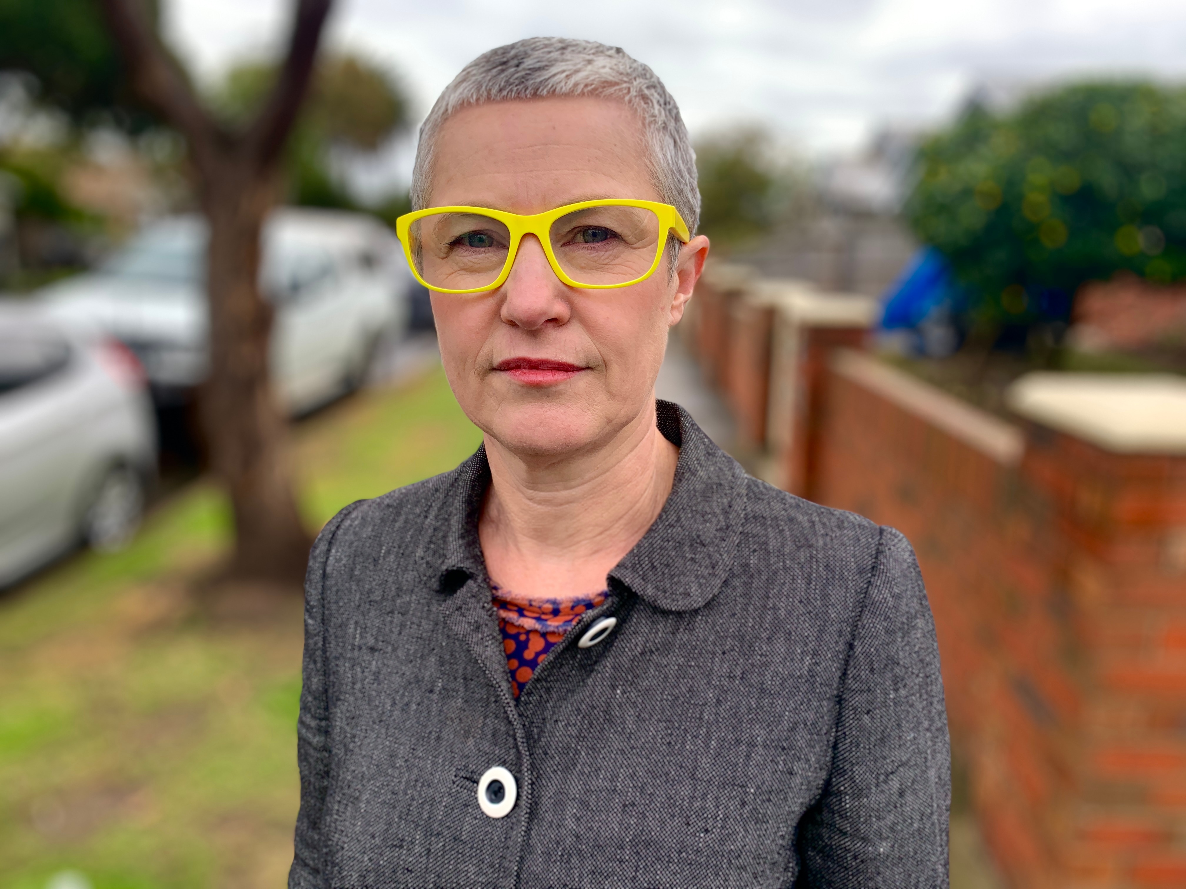 A woman with with a serious expression, short grey hair and yellow glasses stands on a suburban street.