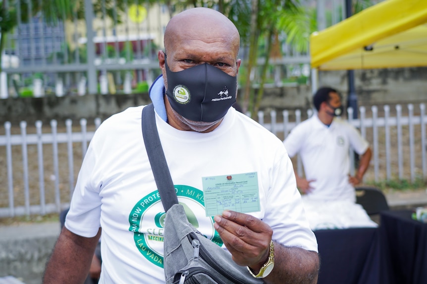 A Papua New Guinean man in a black face mask holds up his vaccination card