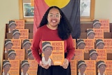 A woman with long dark hair holding an orange book with an aboriginal flag draped in background