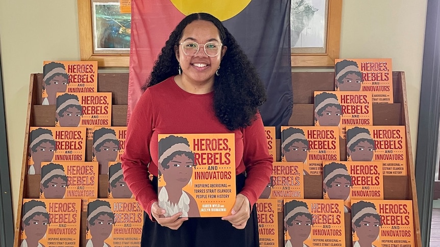 A woman with long dark hair holding an orange book with an aboriginal flag draped in background