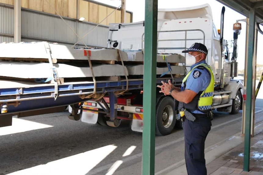 A police officer wearing a mask stands under a shed-like structure next to a truck.